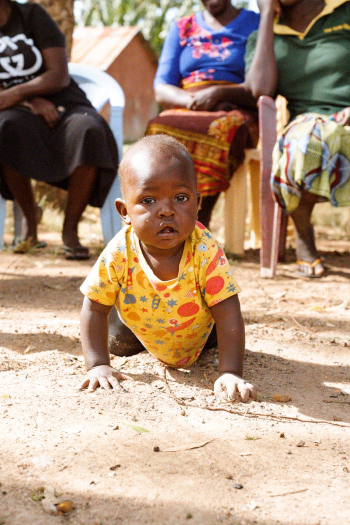 A baby crawls on sandy ground outdoors with seated adults, capturing a moment of exploration.
